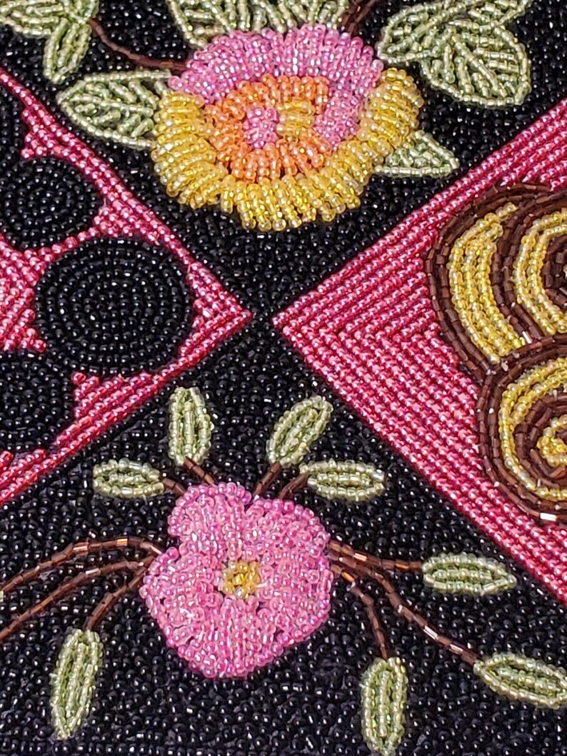 May include: Close-up of a beaded fabric with a floral design. The fabric features pink, yellow, and green beads arranged in a pattern of flowers and leaves. The background is black.