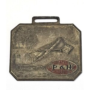 May include: A silver-toned metal watch fob with a raised image of a construction crane and the text "Quality P & H Service".