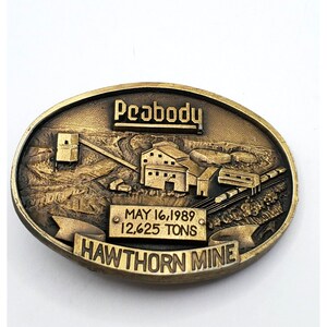 May include: A gold-toned belt buckle with a raised image of a mine and the text "Peabody Hawthorn Mine" and "May 16, 1989 12,625 Tons".