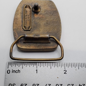Op de afbeelding: Een messing gesp met een reli&euml;fdesign. De gesp is 5 cm breed en heeft een 2MD-markering op de achterkant. De achterkant van de gesp heeft ook de tekst "Indiana Metal Craft &copy; 1977" en het nummer "H95".