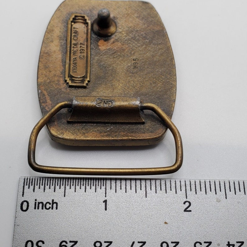 Op de afbeelding: Een messing gesp met een reli&euml;fdesign. De gesp is 5 cm breed en heeft een 2MD-markering op de achterkant. De achterkant van de gesp heeft ook de tekst "Indiana Metal Craft &copy; 1977" en het nummer "H95".