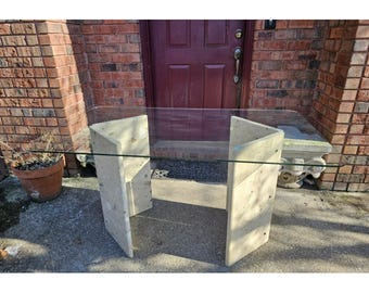 May include: A glass-top table with a rectangular shape and two beige stone bases. The table is in front of a brick wall with a wooden door.