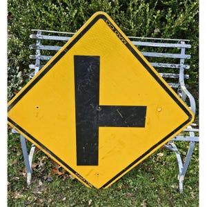 May include: A yellow and black road sign with a T-intersection symbol. The sign is diamond-shaped with the text "MAD CO" along the edges. The sign is resting on a metal bench in front of green foliage.