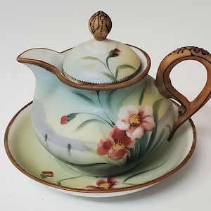 May include: A delicate porcelain creamer and saucer set with a floral design. The creamer features a lid with a knob and a brown handle. The saucer has a gold rim.