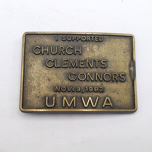May include: A rectangular, bronze-colored metal plaque with raised lettering. The text reads "I SUPPORTED CHURCH CLEMENTS CONNORS NOV.9,1982 UMWA". The plaque has a slightly aged appearance, with some surface wear.