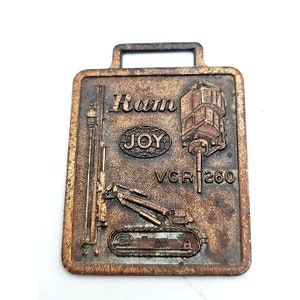 May include: A bronze-colored metal watch fob with a raised image of a drilling rig. The fob features the text "Ram Joy VCR 260".