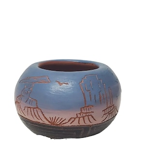 May include: A small, round, blue and brown ceramic pot with a carved design of a desert landscape. The pot is hand-painted and has a rustic, southwestern style.