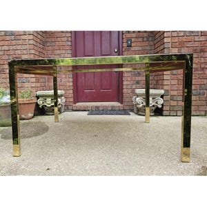 May include: A rectangular brass table with a reflective surface. The table has a simple, geometric design with four square legs. The legs are capped with brass at the bottom. The table is outdoors, in front of a brick wall and a maroon door.