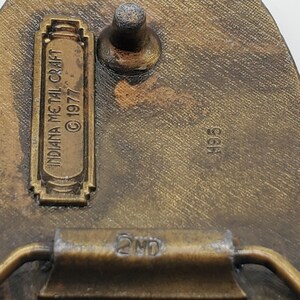 Op de afbeelding: Close-up van een messing gesp met de tekst "INDIANA METAL CRAFT &copy; 1977" en "4955" op de achterkant. De gesp heeft een 2nd markering aan de zijkant.