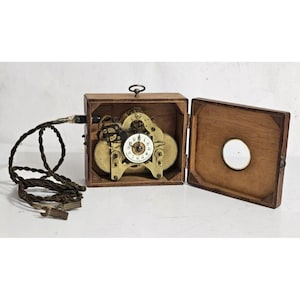 May include: A vintage wooden box with a brass clock mechanism inside. The box is open and the clock face is visible. The clock has a white face with black numbers and hands. The box is made of dark wood and has a brass latch.