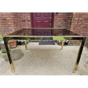 May include: A rectangular glass-top table with a gold-colored metal frame. The table has a sleek, modern design with a reflective surface. The legs are square and angled, providing stability. The table is approximately 60 inches long and 30 inches wide.