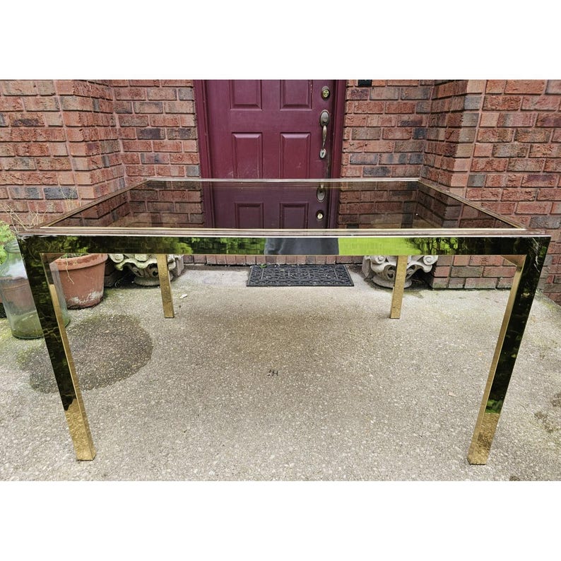 May include: A rectangular glass-top table with a gold-colored metal frame. The table has a sleek, modern design with a reflective surface. The legs are square and angled, providing stability. The table is approximately 60 inches long and 30 inches wide.