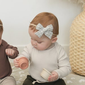 Könnte beinhalten: Zwei Babys in braunen und beigen Leggings und langärmeligen Shirts spielen mit bunten Stapelbechern auf einer weißen und grauen gemusterten Decke. Die Babys tragen passende Stirnbänder mit Schleifen.