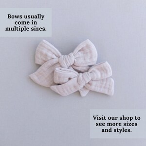 May include: Two beige fabric hair bows of different sizes. The bows are tied together in the center. The text "Bows usually come in multiple sizes. Visit our shop to see more sizes and styles." is printed below the bows.