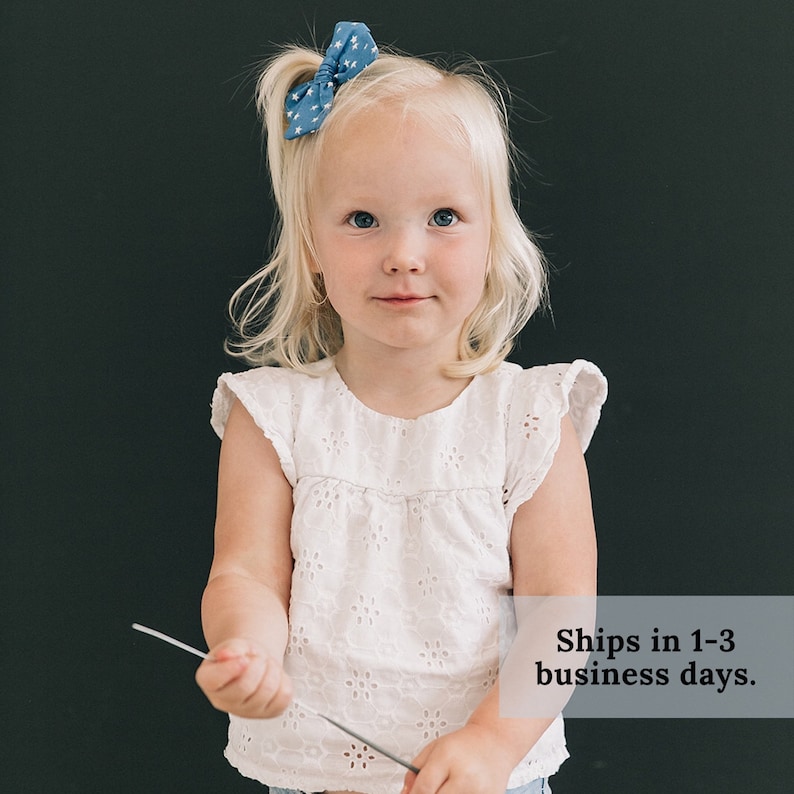 May include: A young girl with blonde hair wearing a white eyelet top and a blue star patterned hair bow. She is holding a silver sparkler in her hand.