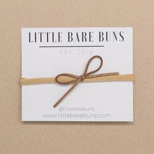 May include: A tan elastic headband with a brown suede bow. The headband is on a white card with the text "LITTLE BARE BUNS EST. 2018 @littlebarebuns www.littlebarebuns.com"
