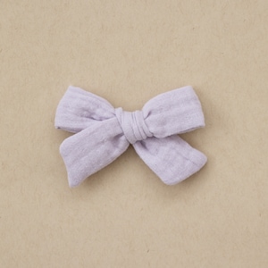 May include: A lavender fabric bow with a simple, tied design. The bow is made of a soft, textured fabric.