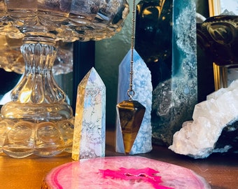 May include: A pink agate coaster with a gold rim and a clear crystal pendulum hanging above it. The pendulum is attached to a chain and has a gold-colored tip. There are several other crystals in the background, including a clear crystal, a blue crystal, and a white crystal.
