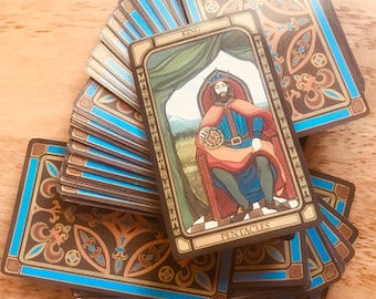 May include: A deck of tarot cards featuring a card with the word "KING" and "PENTACLES". The cards have a blue border and ornate designs. The King card depicts a seated figure in a red robe, holding a coin.