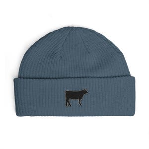 May include: A slate blue ribbed knit beanie featuring a small, embroidered black silhouette of a cow centered on the front cuff. The hat has a thick, folded brim and a textured, vertical knit pattern throughout the entire garment.