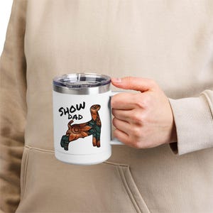Può includere: Tazza da viaggio bianca con bordo e manico argentati. La tazza presenta un'illustrazione decorativa di un cane con un motivo floreale nei toni dell'arancione, del verde e del marrone. Le parole "SHOW DAD" sono stampate in nero sopra il cane.