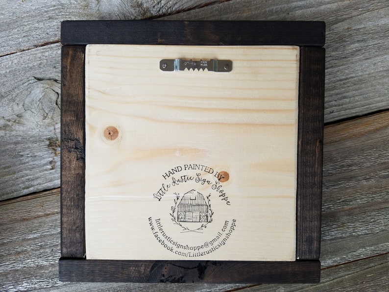 May include: The back of a wooden sign with a dark brown frame. The sign has a black stamp that says "Hand Painted by Little Rustic Sign Shoppe" with a website and email address.