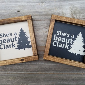 May include: Two wooden framed signs with the text "She's a beaut Clark" and a silhouette of a pine tree. One sign has a white background with black text and tree, the other has a black background with white text and tree.