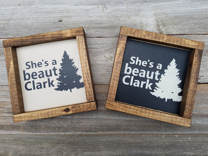 May include: Two wooden framed signs with the text "She's a beaut Clark" and a silhouette of a pine tree. One sign has a white background with black text and tree, the other has a black background with white text and tree.