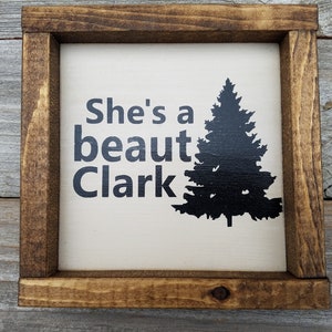 May include: A white wooden sign with a brown frame. The sign has black text that reads "She's a beaut Clark" and a black silhouette of a pine tree.