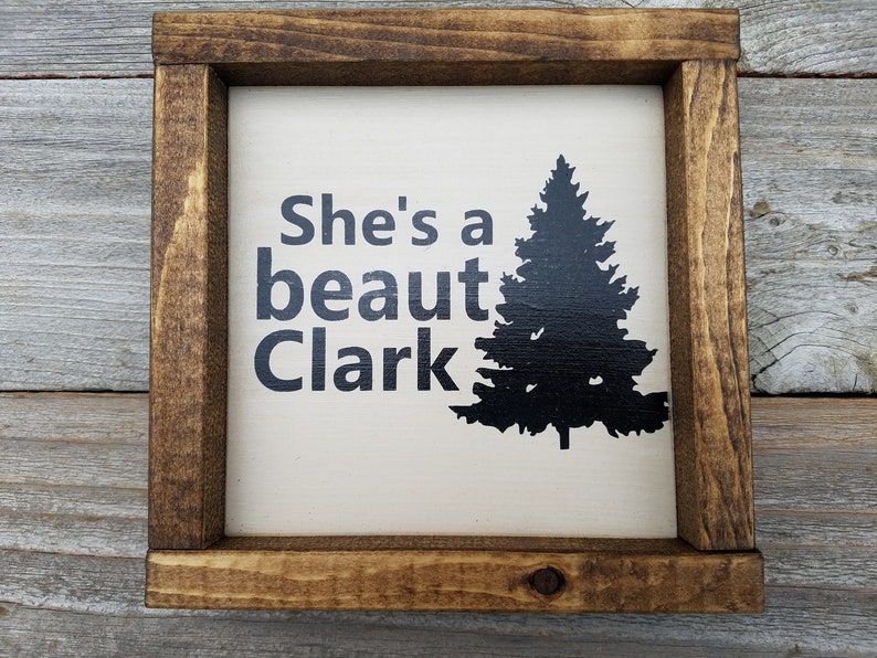 May include: A white wooden sign with a brown frame. The sign has black text that reads "She's a beaut Clark" and a black silhouette of a pine tree.