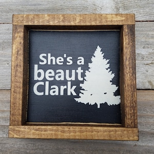 May include: A black wooden sign with a white silhouette of a pine tree and the text "She's a beaut Clark" in white.
