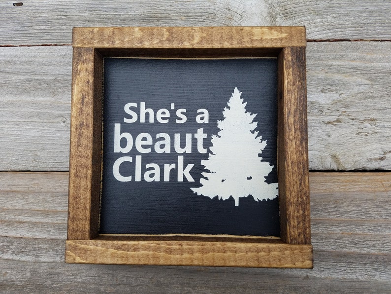 May include: A black wooden sign with a white silhouette of a pine tree and the text "She's a beaut Clark" in white.