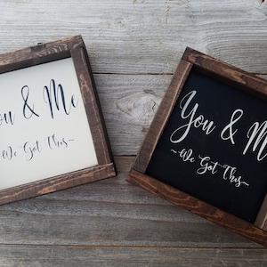 May include: Two wooden framed signs with the text "You & Me - We Got This" in black lettering on a white background and white lettering on a black background.
