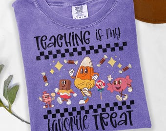 May include: Violet Comfort Colors t-shirt with the text "Teaching is my favorite treat." The shirt features a candy corn character, chocolate bar, and other candy graphics. The shirt has a crew neck and short sleeves.