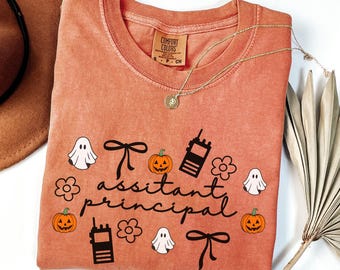 May include: A yam-colored Comfort Colors crewneck sweatshirt with the words "assistant principal" and Halloween-themed graphics. The design includes ghosts, pumpkins, bows, and walkie-talkies. A gold necklace with a pendant is draped on the shirt.