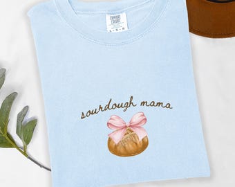 May include: Light blue t-shirt with the text "sourdough mama" and a cartoon illustration of a sourdough bread loaf with a pink ribbon. The shirt is made of a soft, comfortable chambray fabric.