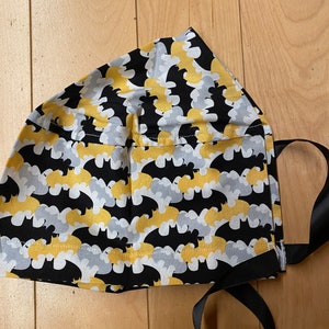 Puede incluir: Un gorro de cocinero de tela negro, amarillo y blanco con un patrón de murciélago. El gorro tiene cintas de raso negras.