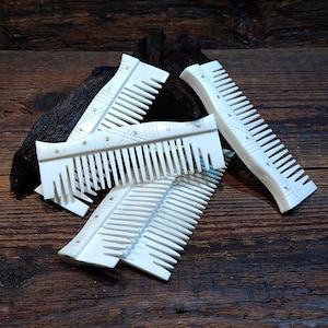 May include: Four white bone combs with intricate designs on a wooden surface. The combs are arranged in a pile, with the teeth facing up. The combs are made of bone and have a natural, polished finish.