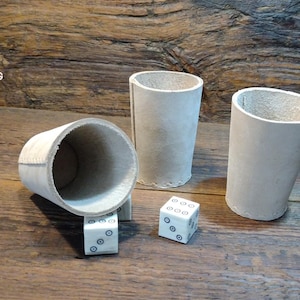 Può includere: Tre tazze cilindriche di colore chiaro e due dadi bianchi con punti blu sono disposti su una superficie di legno. Le tazze sono realizzate in un materiale strutturato e i dadi sono piccoli e quadrati. Il logo Viking Thunder è in alto a sinistra.