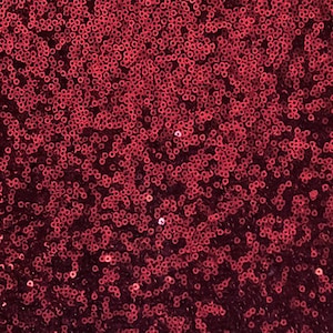 May include: A close-up shot of a fabric covered in small, round, shiny, red sequins. The sequins are densely packed, creating a textured surface. The colour transitions from a lighter red at the top to a darker, burgundy shade at the bottom.