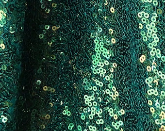 May include: Close-up of a shimmering emerald green fabric covered in small, round sequins. The sequins catch the light, creating a sparkling effect. The fabric has a textured, slightly rippled appearance, suggesting a flowing material.