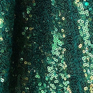 Op de afbeelding: Close-up van een glinsterende smaragdgroene stof bedekt met kleine, ronde pailletten. De pailletten vangen het licht op en creëren een sprankelend effect. De stof heeft een getextureerde, licht gerimpelde uitstraling.