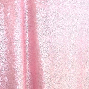 Op de afbeelding: Een close-up van een roze paillettenstof. De stof is lichtroze van kleur en is bedekt met kleine, ronde pailletten die het licht vangen. De pailletten zijn dicht op elkaar gepakt, waardoor een glinsterend effect ontstaat. De stof wordt waarschijnlijk gebruikt voor kleding of decoratieve doeleinden.