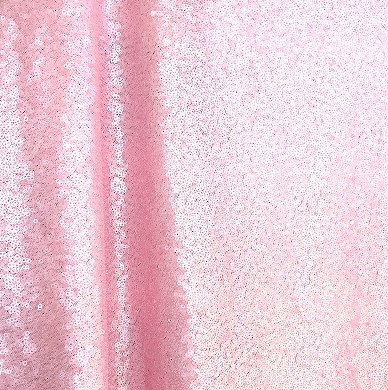 Op de afbeelding: Een close-up van een roze paillettenstof. De stof is lichtroze van kleur en is bedekt met kleine, ronde pailletten die het licht vangen. De pailletten zijn dicht op elkaar gepakt, waardoor een glinsterend effect ontstaat. De stof wordt waarschijnlijk gebruikt voor kleding of decoratieve doeleinden.