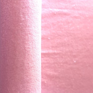 Op de afbeelding: Een rol roze stof bedekt met kleine, ronde pailletten. De stof is strak opgerold aan de linkerkant van de afbeelding, met de uitgerolde stof zichtbaar aan de rechterkant. De pailletten vangen het licht en cre&euml;ren een glinsterend effect.