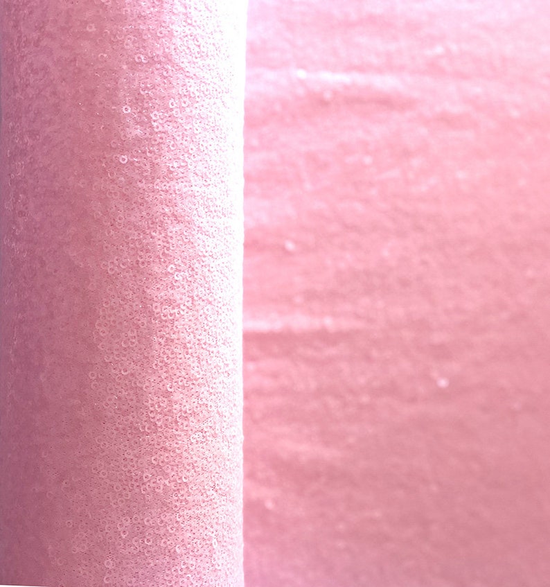 Op de afbeelding: Een rol roze stof bedekt met kleine, ronde pailletten. De stof is strak opgerold aan de linkerkant van de afbeelding, met de uitgerolde stof zichtbaar aan de rechterkant. De pailletten vangen het licht en cre&euml;ren een glinsterend effect.