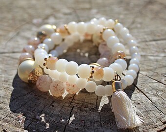 Puede incluir: Un brazalete de capas con cuentas blancas y rosadas, detalles dorados y un borla blanca.