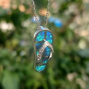 Op de afbeelding: Een zilveren ketting met een blauwe opaal flip-flop hanger. De hanger is versierd met kleine zilveren accenten.