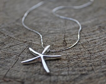 May include: A silver chain necklace with a silver starfish pendant. The necklace is lying on a piece of wood.