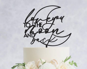 May include: A two-tiered white cake decorated with white flowers and greenery. A black cake topper in the shape of a crescent moon with the words "love you to the moon and back" is placed on top. The cake sits on a white cake stand.